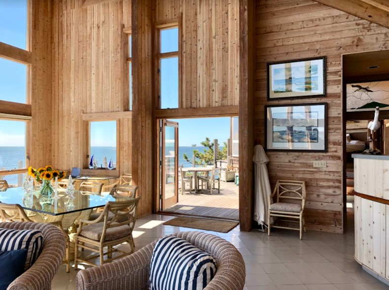 Interior view of a wooden house featuring a dining table with flowers, a cozy seating area, and large windows offering a scenic view of the ocean.