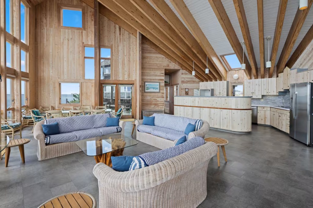 Interior view of a spacious living area featuring wooden accents, large windows, and comfortable seating. The kitchen is visible in the background with modern appliances.