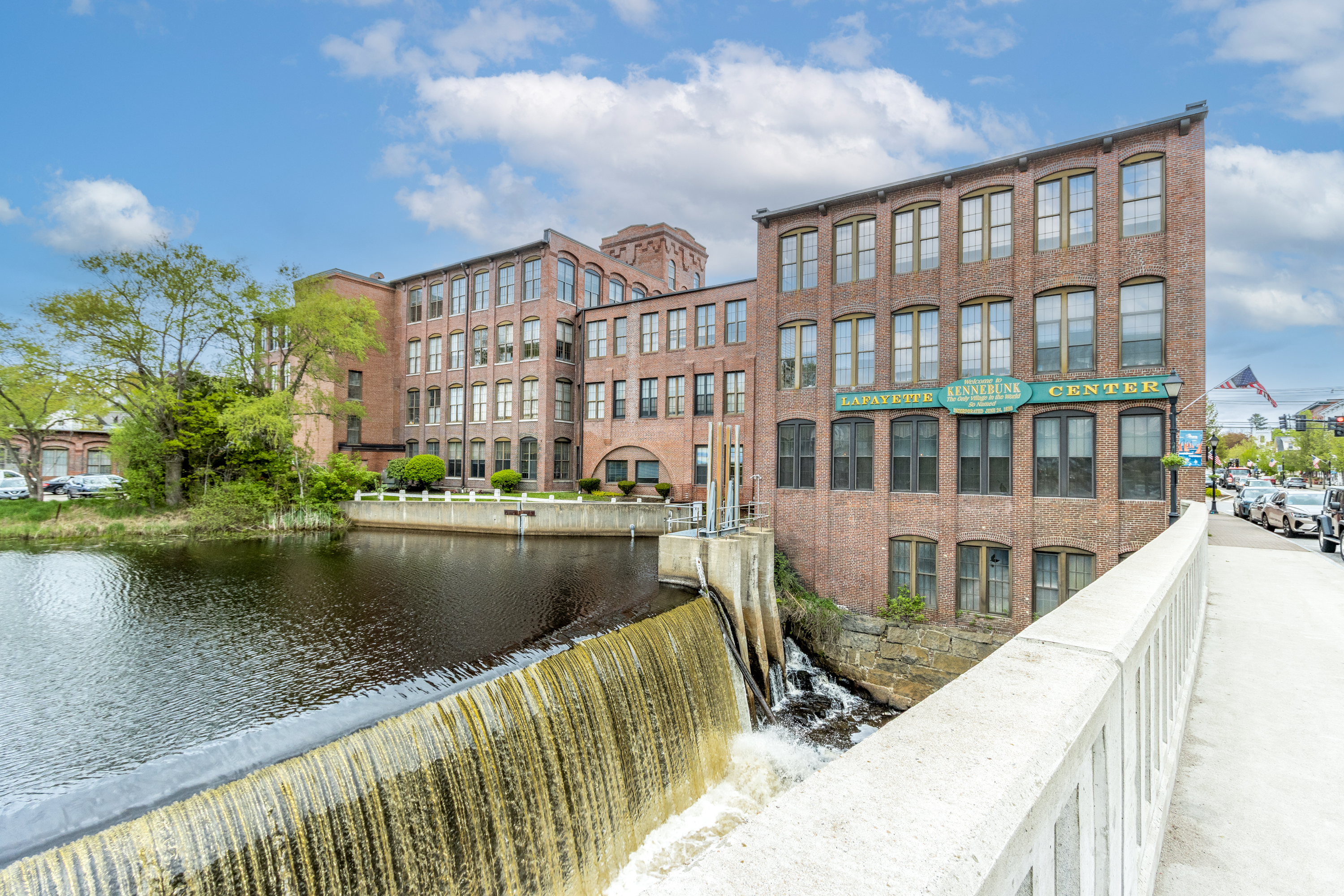 View of the Lafayette Center building in Kennebunk, Maine, featuring a dam with water cascading down, surrounded by greenery and urban elements.