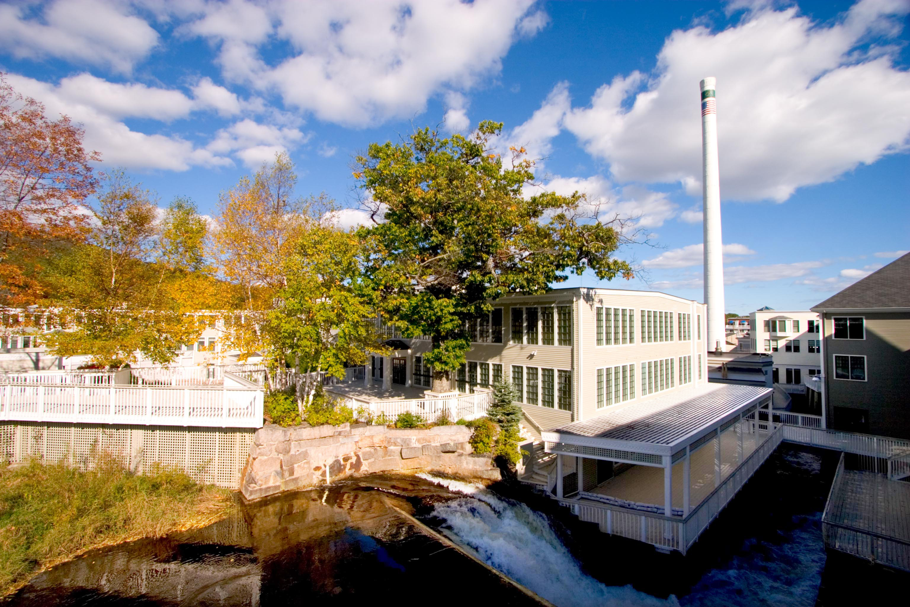 A view of a riverside building complex with large windows, surrounded by autumn foliage and a tall smokestack, under a partly cloudy sky.