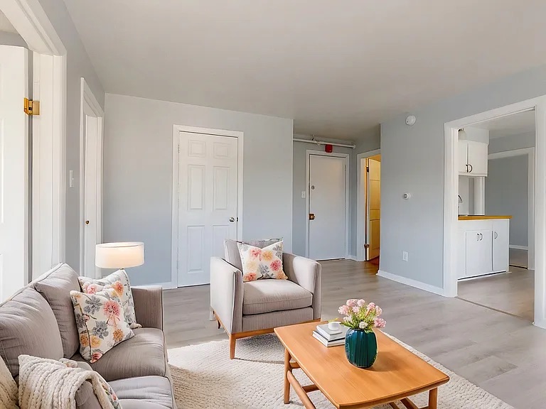 A cozy living room featuring a light gray wall, a gray sofa adorned with floral cushions, a light wood coffee table with a green vase and flowers, and a warm lamp beside the sofa. Multiple doors and a small kitchenette are visible in the background.